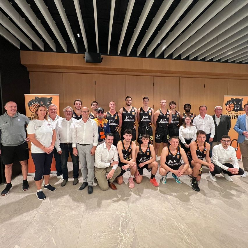 Ein Basketballteam posiert mit Offiziellen und Unterstützern in einem Konferenzraum mit Holzwand und Deckenbalken.