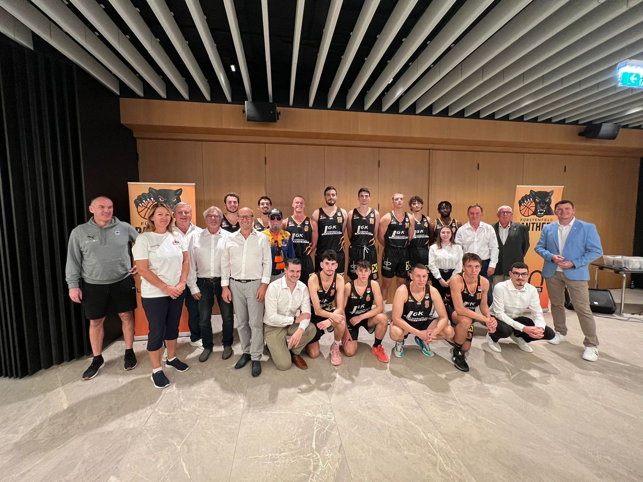 Ein Basketballteam posiert mit Offiziellen und Unterstützern in einem Konferenzraum mit Holzwand und Deckenbalken.