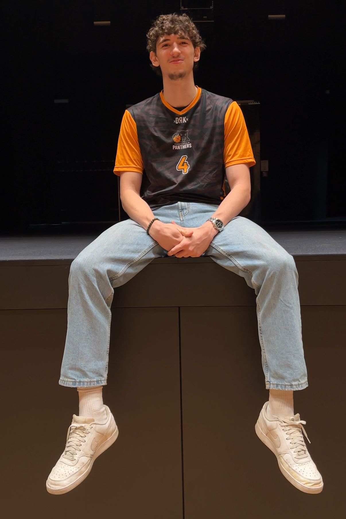Ein Mann sitzt auf einer Bühne und trägt ein Panthers-Trikot mit der Nummer 4. Er trägt blaue Jeans und weiße Turnschuhe.