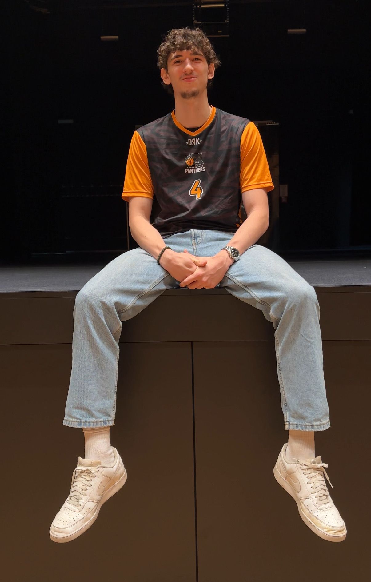Ein Mann sitzt auf einer Bühne und trägt ein Panthers-Trikot mit der Nummer 4. Er trägt blaue Jeans und weiße Turnschuhe.