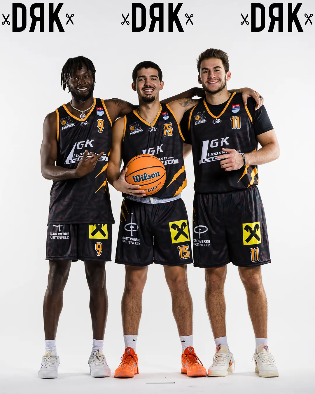 Drei Basketballspieler in schwarzen Trikots mit gelben Akzenten und den Nummern 9, 15 und 11 posieren für ein Foto. Der Spieler in der Mitte hält einen Wilson-Basketball.