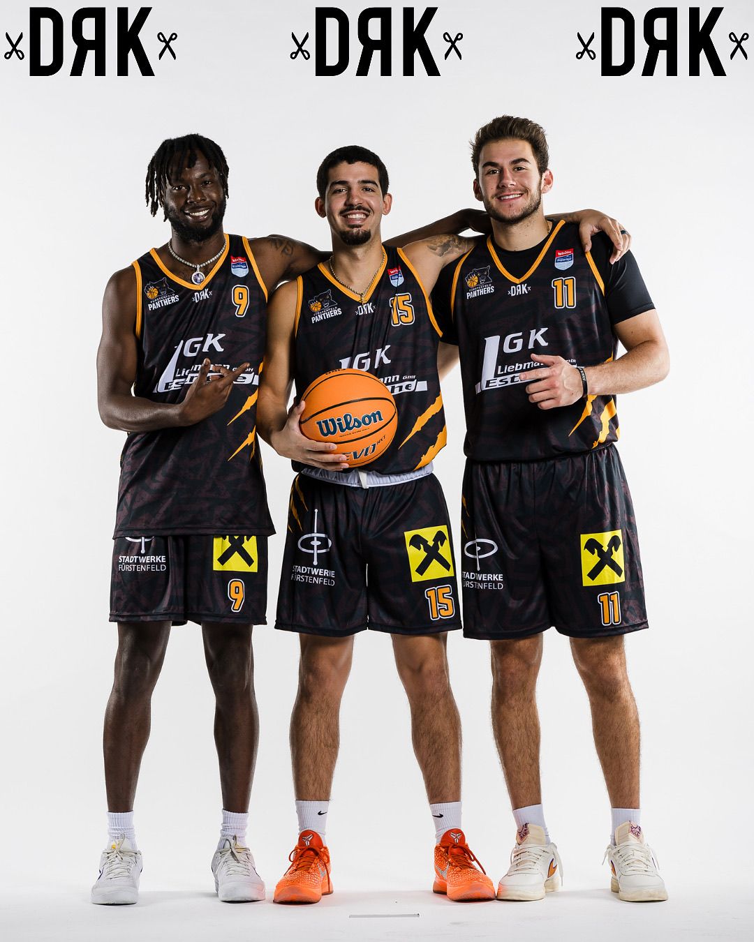 Drei Basketballspieler in schwarzen Trikots mit gelben Akzenten und den Nummern 9, 15 und 11 posieren für ein Foto. Der Spieler in der Mitte hält einen Wilson-Basketball.