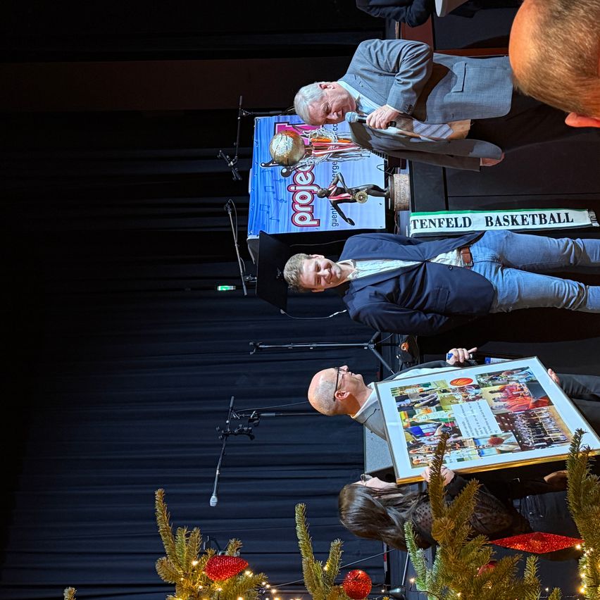 Eine Gruppe von Männern und einer Frau auf einer Bühne während einer Preisverleihung, einer hält eine gerahmte Collage mit Basketball-Thematik. Der Hintergrund zeigt ein Tenfeld Basketball-Banner.