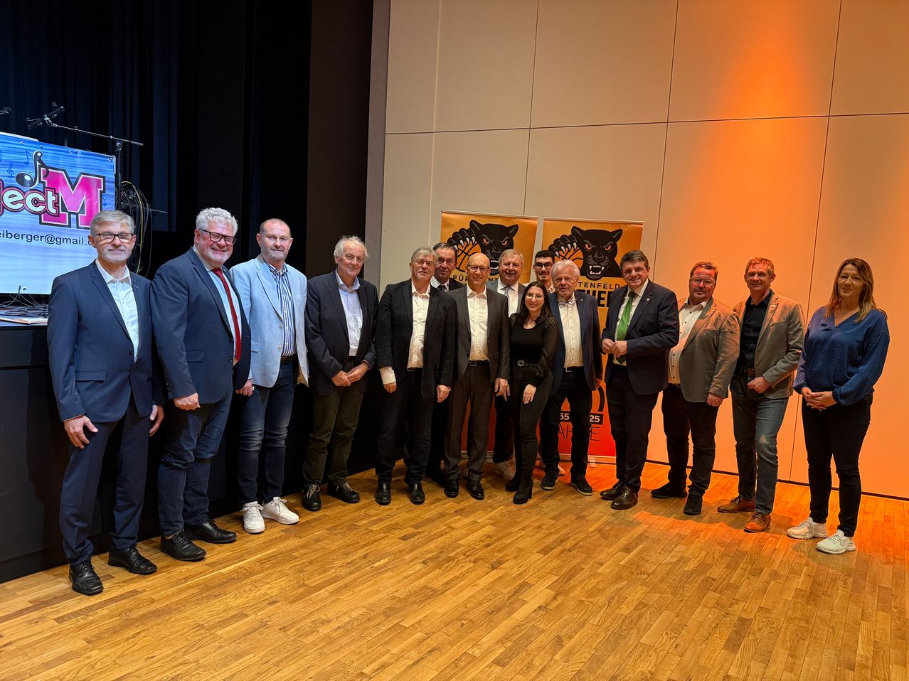 Eine Gruppe formell gekleideter Personen, einschließlich Männer und Frauen, posiert für ein Foto vor Bannern mit einem schwarzen Panther-Logo.