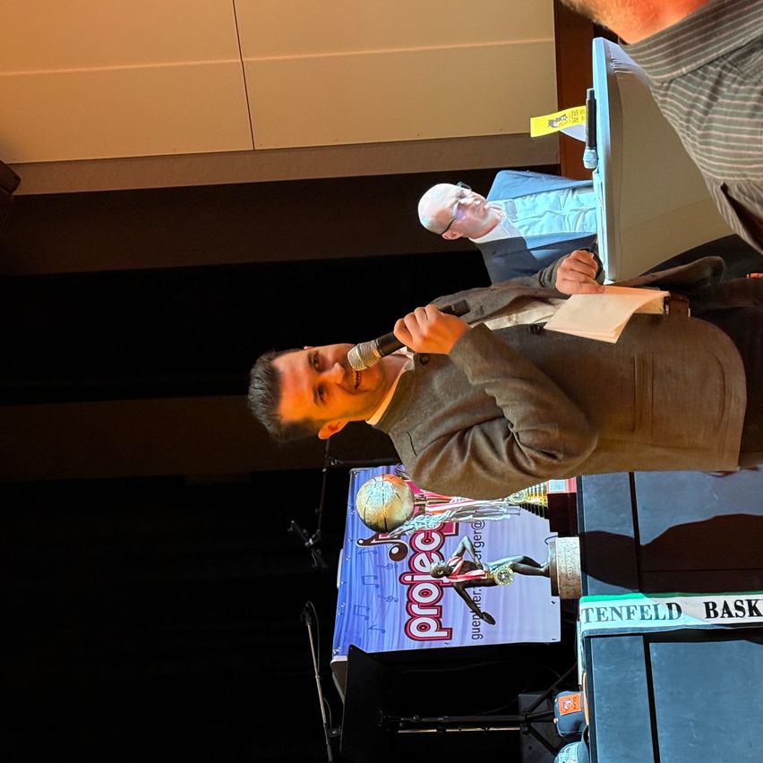 Ein Mann spricht in ein Mikrofon auf der Bühne. Hinter ihm hält eine Person ein Papier. Ein Banner liest Project und Tenfeld Basket.