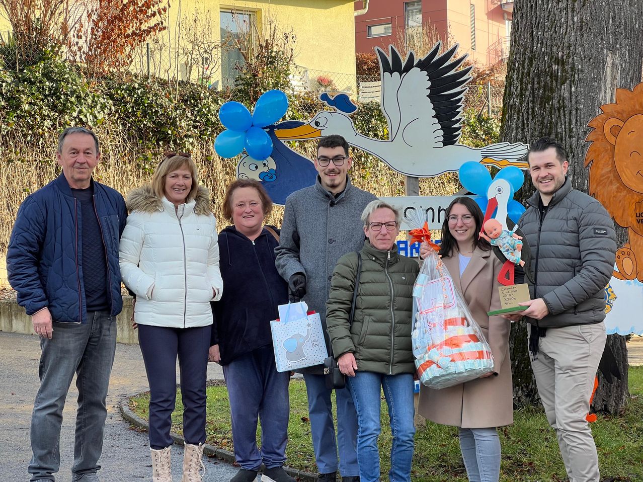 Eine Gruppe von sechs Erwachsenen, zwei Frauen und vier Männer, stehen zusammen vor einem Storch-Schild und halten Geschenktüten und eine Puppe.