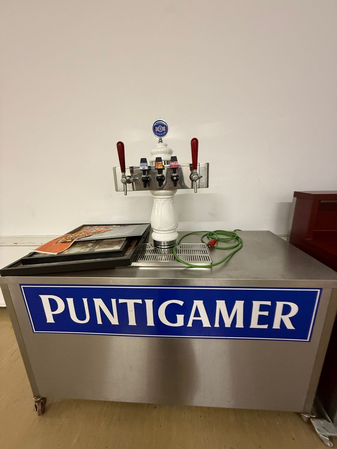 Ein Edelstahlspender mit einem blauen Schild, auf dem 'Puntigamer' steht. Es hat rote Griffe und mehrere Zapfhähne, die mit einem grünen Kabel verbunden sind.