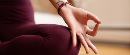 Eine Frau in Weinrothosen praktiziert Yoga, ihre Hand in einer meditativen Geste. Sie hat ein Tattoo am Arm.