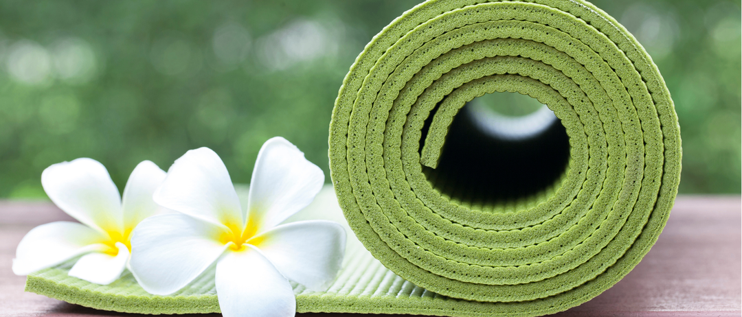 Eine aufgerollte grüne Yogamatte liegt auf einer Holzoberfläche, mit weißen Blumen und einem unscharfen grünen Hintergrund.