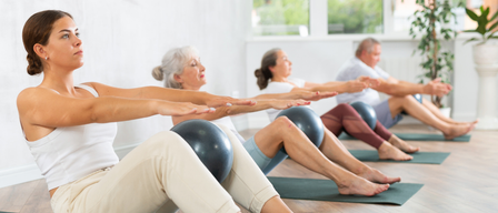 Four people on yoga mats are performing seated exercises. Each person holds a ball, with three in the foreground and one behind. The room has large windows.
