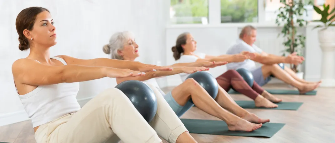 Vier Personen auf Yogamatten führen sitzende Übungen durch. Jede Person hält einen Ball, drei im Vordergrund und eine dahinter. Der Raum hat große Fenster.