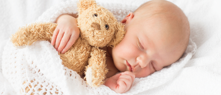 A baby is sleeping on a white blanket, holding a teddy bear in its hand. The baby's eyes are closed.