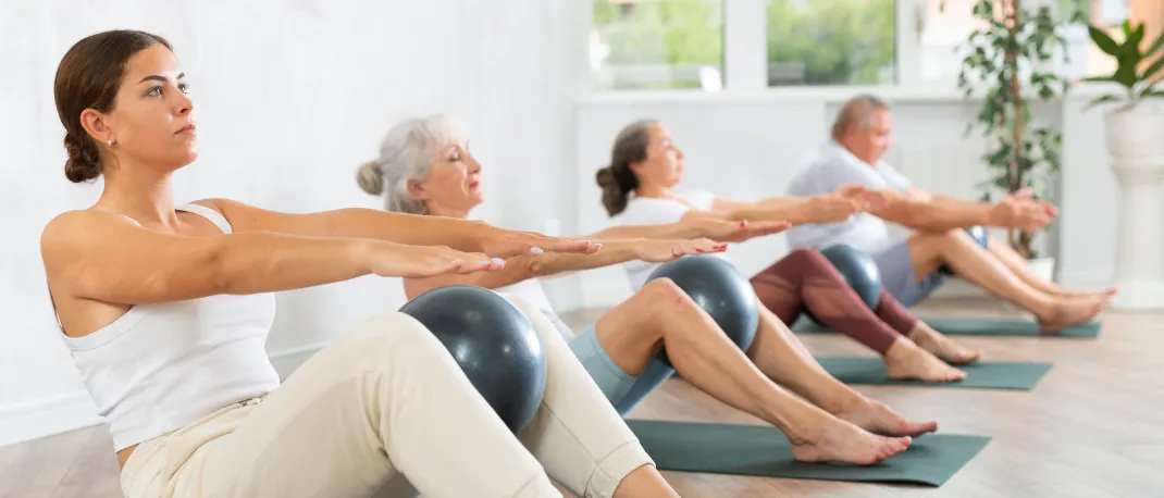 Eine Gruppe von Personen praktiziert Yoga-Übungen auf dem Boden. Eine Frau vorne benutzt einen Yogaball. Eine weitere Frau sitzt hinter ihr, und zwei Männer sitzen hinter ihr.