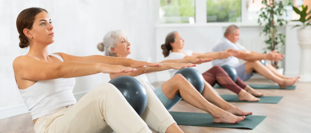 Vier Personen üben auf Yogamatten mit einem Ball, zwei ältere Frauen und ein Mann, alle schauen nach vorne. Dahinter ist eine weiße Wand mit einem Fenster.