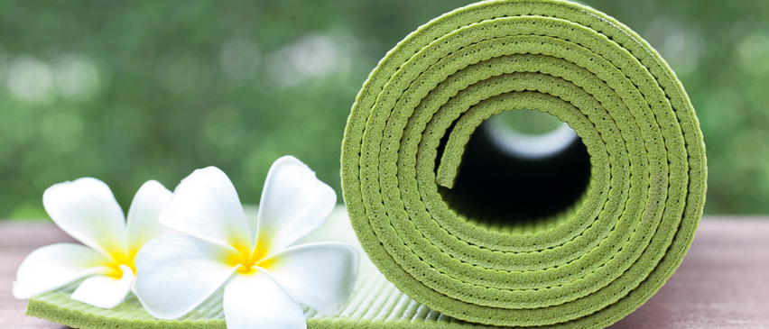 Eine aufgerollte grüne Yogamatte mit weißen Plumeria-Blumen auf einem Holztisch.