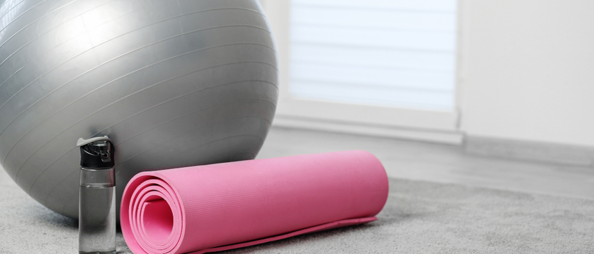 Ein grauer Gymnastikball, eine rosa Yogamatte und eine Wasserflasche liegen auf einem Teppichboden in der Nähe eines Fensters mit Jalousien.