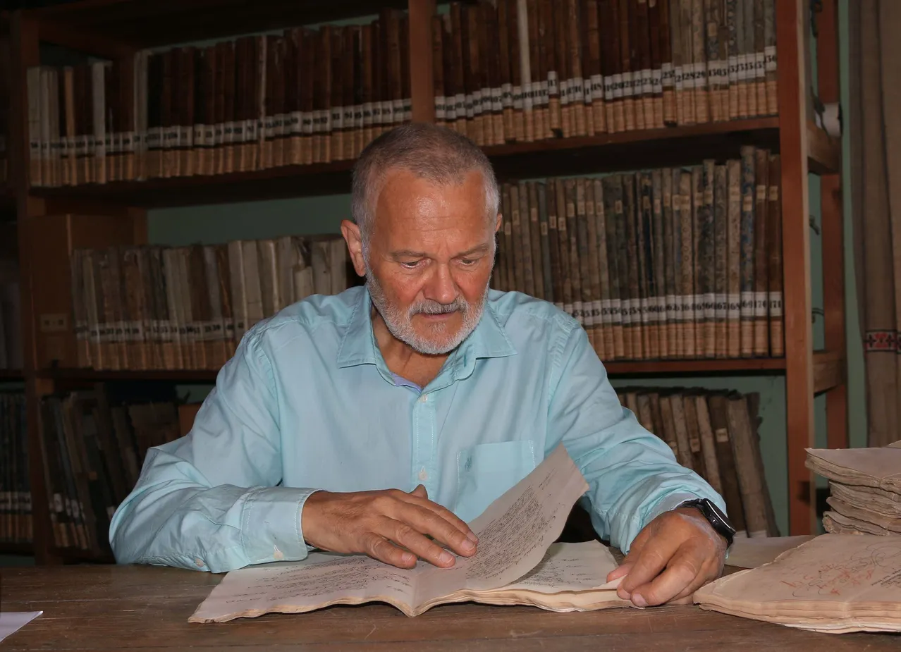 Ein älterer Mann mit Bart in einem blauen Hemd sitzt vor einem Holztisch und liest ein Buch. Im Hintergrund befinden sich Regale mit alten Büchern.