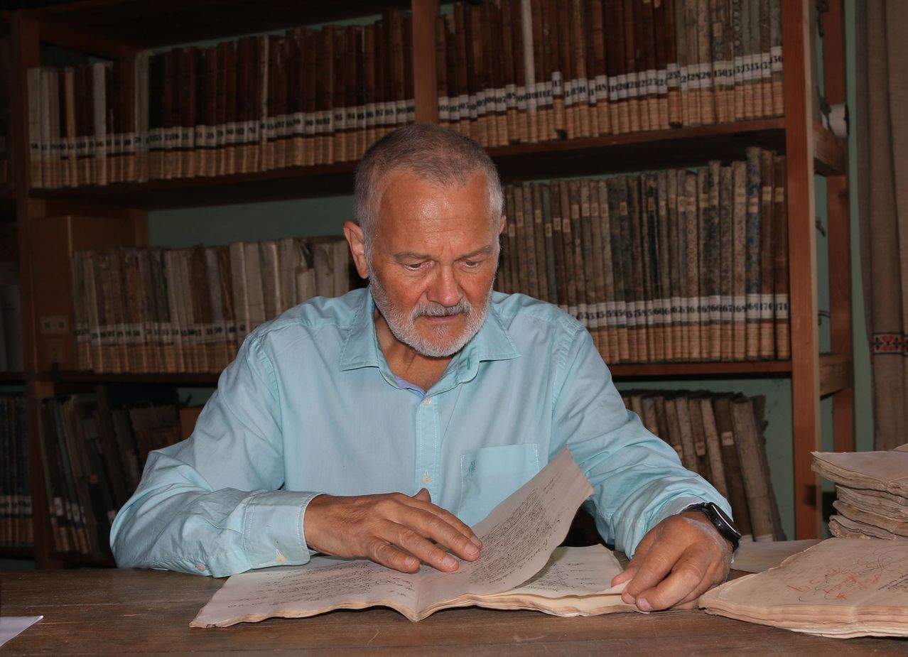 Ein älterer Mann mit Bart in einem blauen Hemd sitzt vor einem Holztisch und liest ein Buch. Im Hintergrund befinden sich Regale mit alten Büchern.