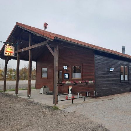 Ein kleines Holzgebäude mit einem braunen Dach und einem Schild, das Gast Haus liest. Das Gebäude hat mehrere Fenster und einen Bildschirm. Es gibt Blumentöpfe draußen.