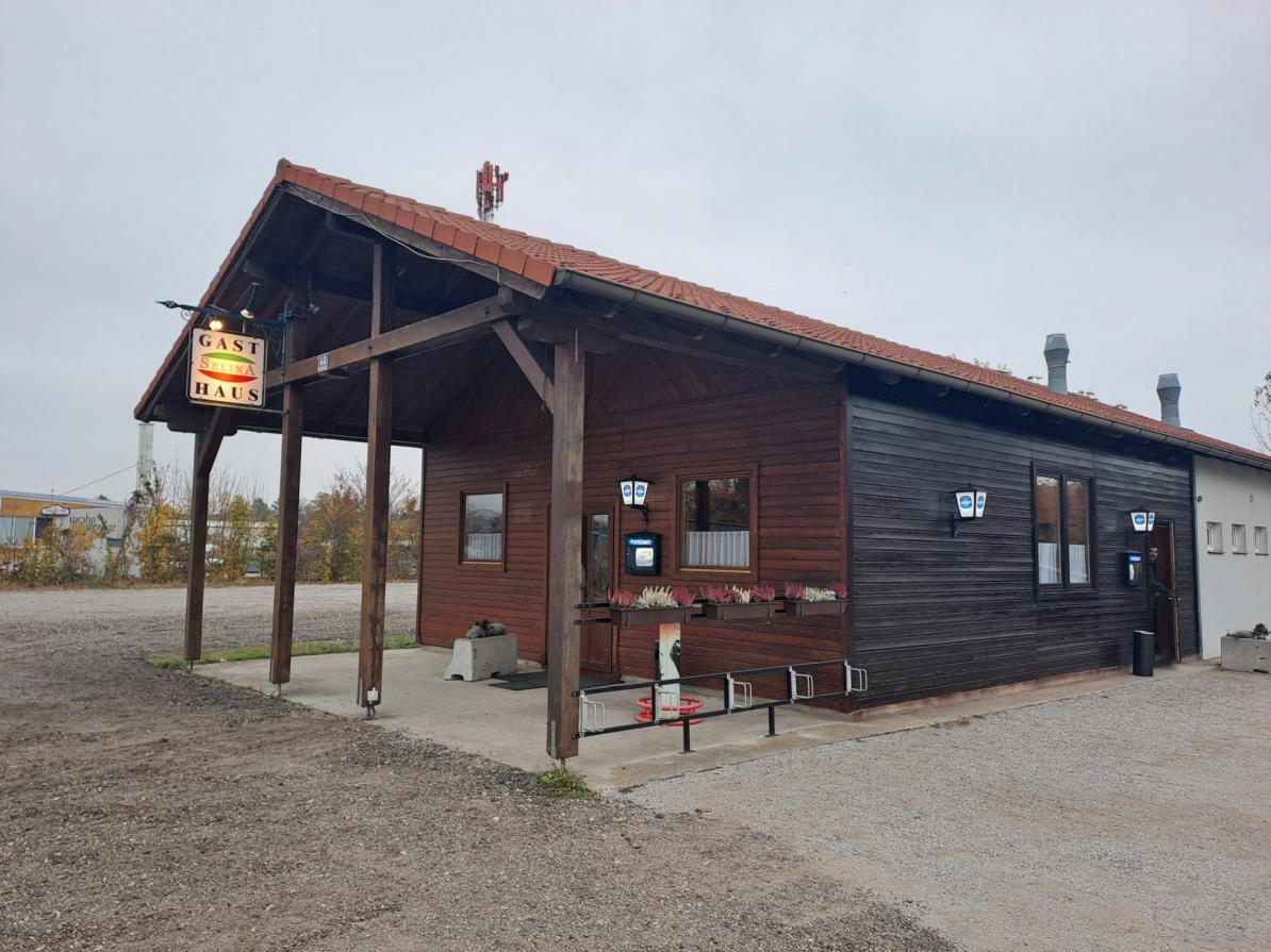 Ein kleines Holzgebäude mit einem braunen Dach und einem Schild, das Gast Haus liest. Das Gebäude hat mehrere Fenster und einen Bildschirm. Es gibt Blumentöpfe draußen.