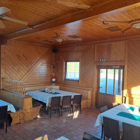 Ein hölzerner Speisesaal mit einem Tisch für Mahlzeiten. Der Raum hat Holzwände und Decken mit Ventilatoren. Es gibt Stühle und Tische mit grünen Tischsets und Gegenständen darauf. Eine Bank mit Geländern steht links und eine Tür mit Glasscheibe rechts. Es gibt ein Fenster mit Blick nach draußen.
