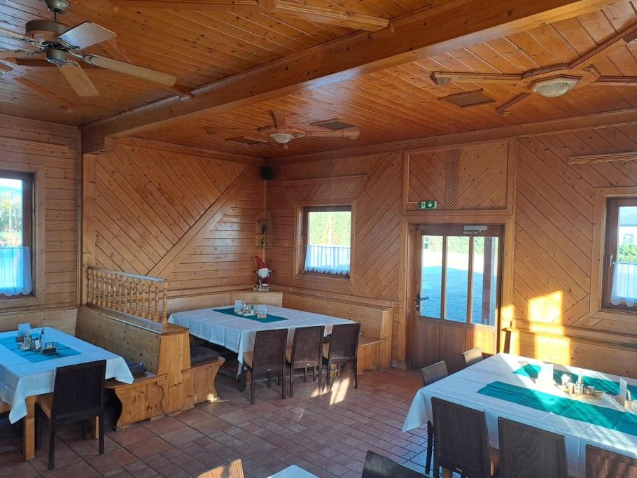 Ein hölzerner Speisesaal mit einem Tisch für Mahlzeiten. Der Raum hat Holzwände und Decken mit Ventilatoren. Es gibt Stühle und Tische mit grünen Tischsets und Gegenständen darauf. Eine Bank mit Geländern steht links und eine Tür mit Glasscheibe rechts. Es gibt ein Fenster mit Blick nach draußen.