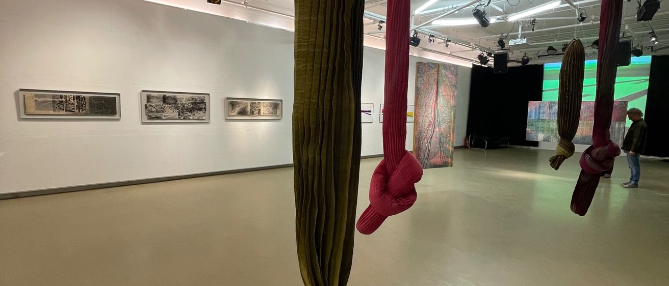 Ein moderner Kunstgalerieraum mit weißen Wänden, der große, farbenfrohe hängende Skulpturen, beleuchtet von Deckenleuchten, mit gerahmten Kunstwerken an der Wand und einer Person, die auf der rechten Seite steht, zeigt.