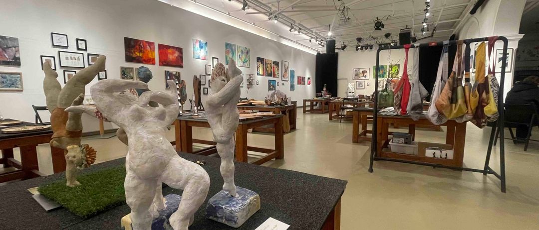 Eine überdachte Galerie mit weißen Wänden und Decken, in der verschiedene Skulpturen und Gemälde ausgestellt sind. Es gibt Holztische mit Skulpturen zur Schau gestellt, zusammen mit anderen Kunstwerken an den Wänden. An der Decke befestigt sind Scheinwerfer.