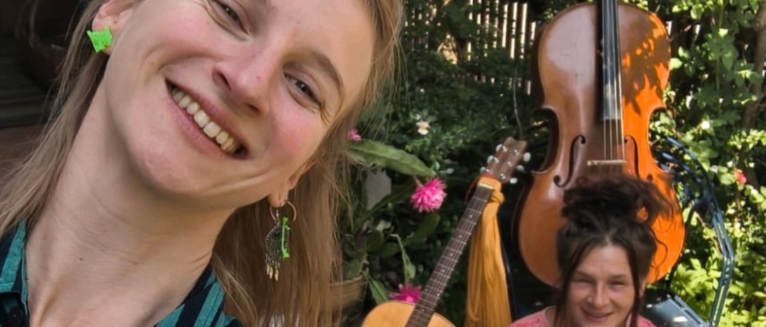 Zwei Frauen sind draußen in einem Garten. Die Frau vorne lächelt und trägt ein gestreiftes Hemd. Hinter ihr sitzt eine weitere Frau auf einem Stuhl mit Musikinstrumenten.