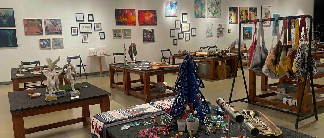 Eine Kunstgalerie mit verschiedenen Kunstwerken an den Wänden, Skulpturen und Schmuck auf Tischen. Verschiedene Gegenstände sind auf Tischen ausgestellt, und Taschen hängen an einem Ständer.