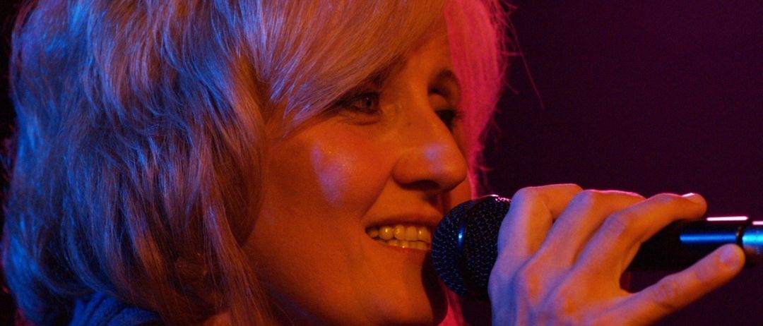 Eine Frau mit kurzen blonden Haaren und einem blauen Pullover singt in ein Mikrofon und lächelt. Sie hat rosa Strähnen und wird von einem lila Licht beleuchtet.