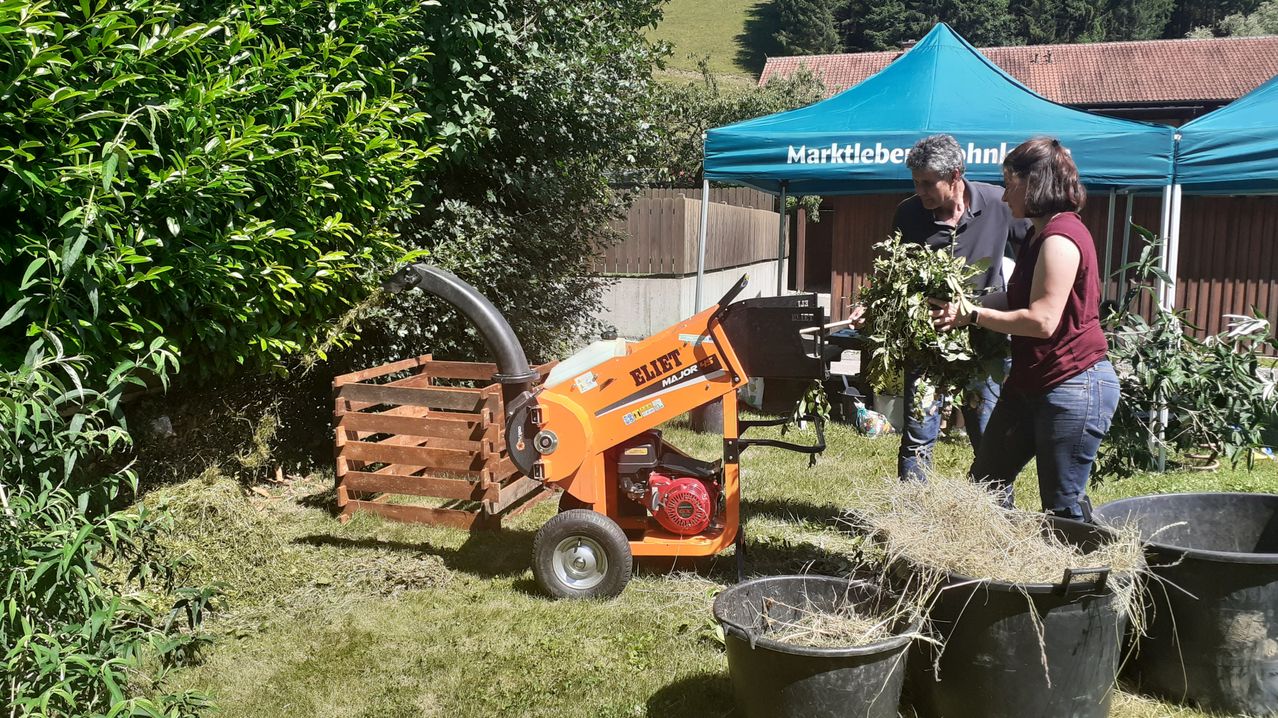 Ein Mann benutzt einen orangefarbenen Laubbläser in einem Garten, mit einem Aufbewahrungskasten und zwei schwarzen Eimern in der Nähe. Er hält eine Handvoll grüner Blätter. Hinter ihm ist ein Zelt mit dem Wort 'Marktleber' und ein Holzzaun zu sehen.