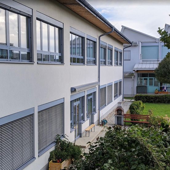 Ein weißes Gebäude mit mehreren Fenstern und geschlossenen Jalousien, einem Garten und einer Bank davor.