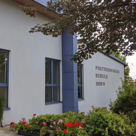 Die Vorderseite eines weißen Gebäudes mit blauen Akzenten. Es steht Polytechnische Schule Horn. Davor befinden sich Pflanzen und Blumen.