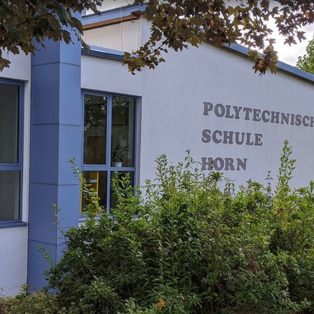 Das Äußere eines Gebäudes mit der Aufschrift Polytechnische Schule Horn an der Wand. Es hat blaue Begrenzungen und ein Fenster mit einer Pflanze darin.