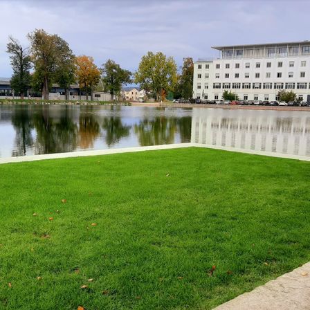 Ein ruhiger Blick auf einen großen, ruhigen See, umgeben von einem üppigen grünen Rasen und verschiedenen Bäumen. Ein modernes weißes Gebäude steht auf der gegenüberliegenden Uferseite mit mehreren geparkten Autos davor.