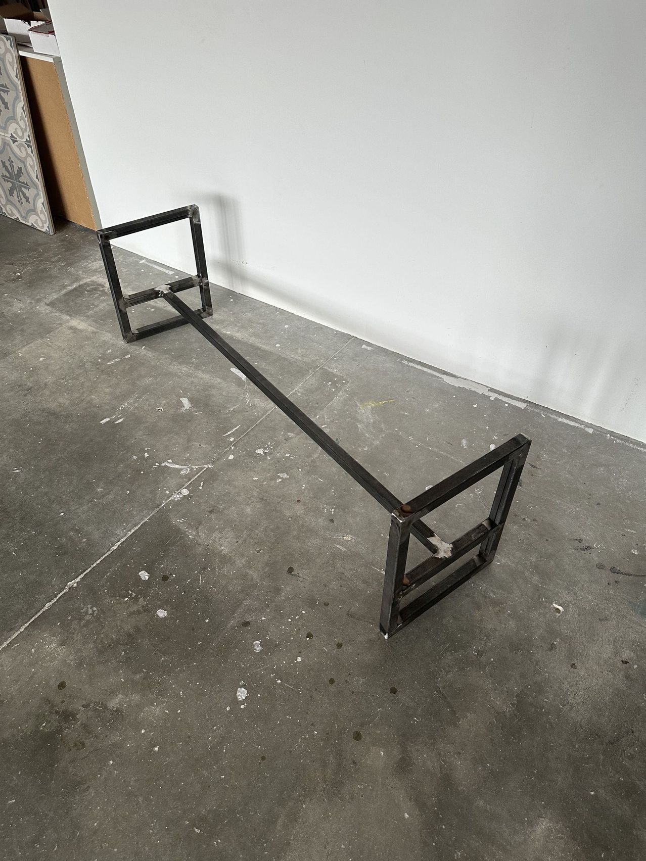 Ein Metall-Bankrahmen liegt auf einem Betonboden mit einer weißen Wand im Hintergrund.