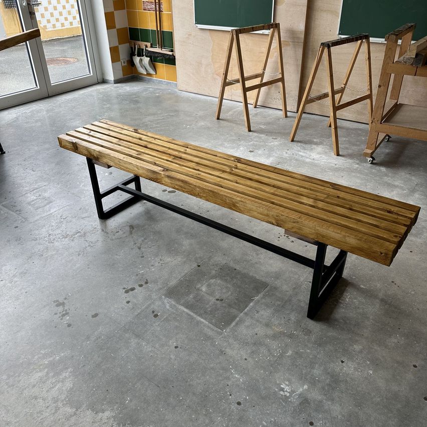 Eine Holzbank mit schwarzen Metallbeinen in einem Raum mit Betonboden und einer Glastür.