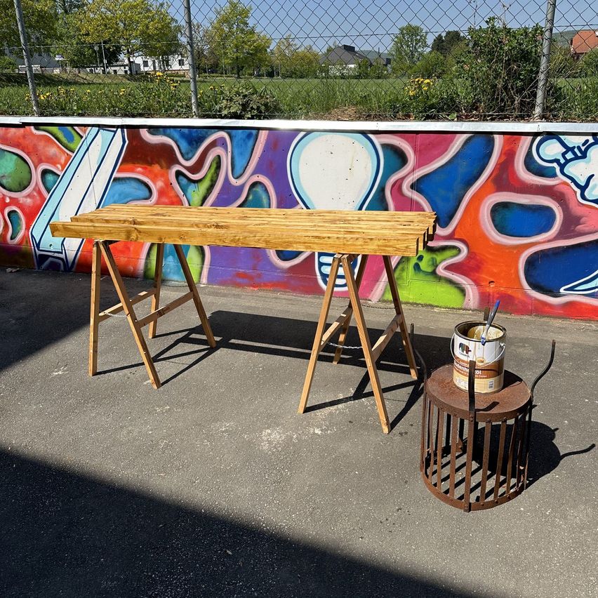 Ein hölzerner Werkbank steht auf einer Betonfläche, mit einem Eimer Farbe auf einem rostigen Käfig neben ihm. Dahinter erstreckt sich eine farbenfrohe, mit Graffiti bedeckte Wand entlang des Zauns.