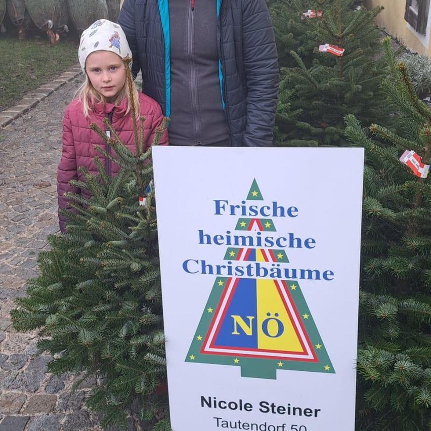 Ein Mädchen und ein Mann stehen neben einem Weihnachtsbaummarkt, mit einem Schild davor, das 'Frische heimische Christbäume' sagt.