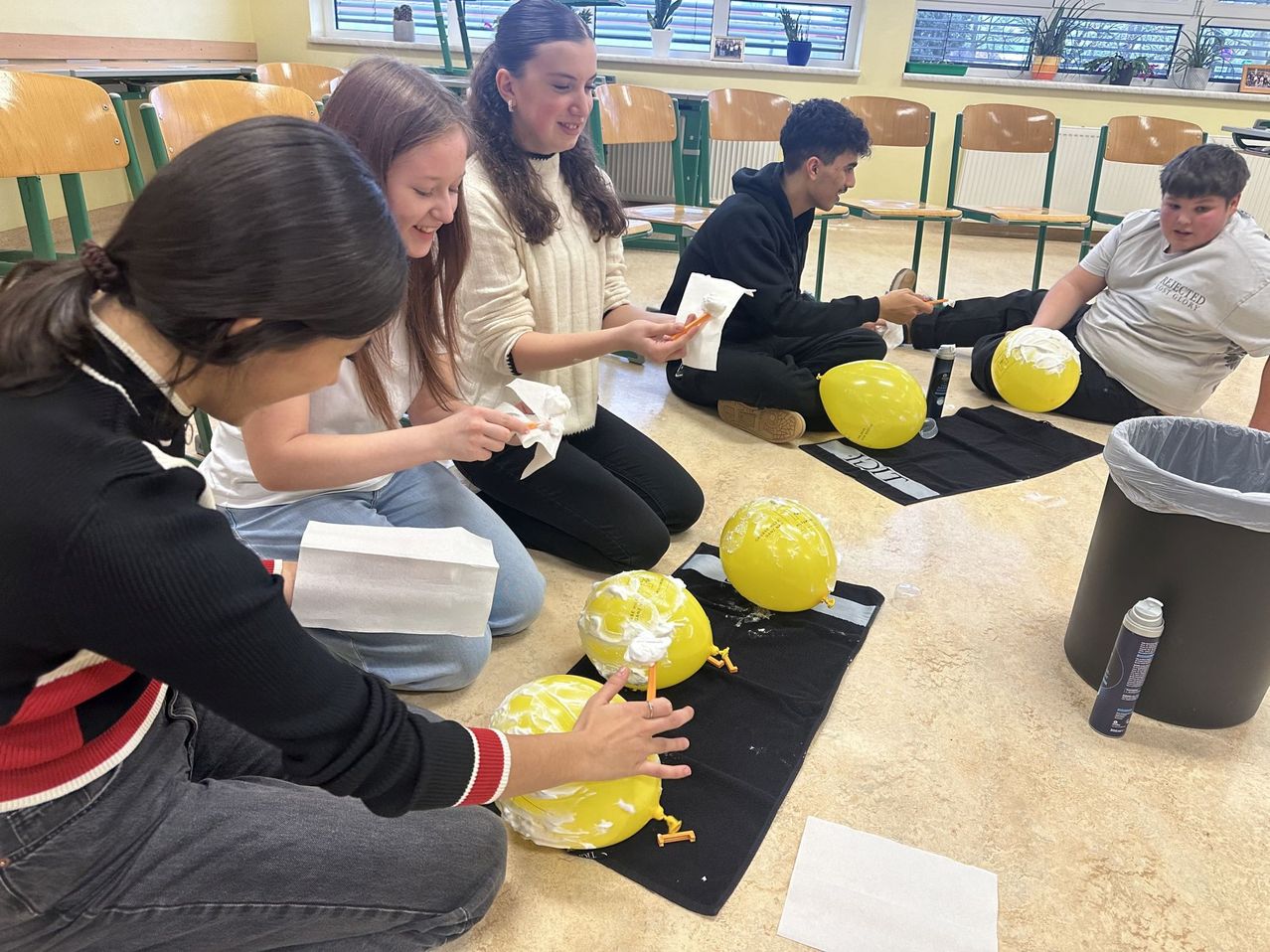 Eine Gruppe von Schülern sitzt auf dem Boden und arbeitet mit gelben Ballons. Sie halten Werkzeuge und scheinen die Ballons zu dekorieren.