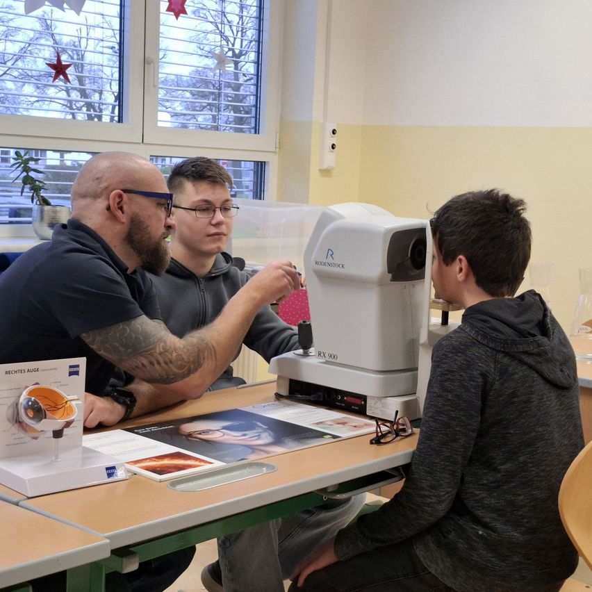 Zwei Männer verwenden ein Augentestgerät an einem jungen Mann in einem Klassenzimmer. Ein Poster eines Auges ist auf dem Tisch.