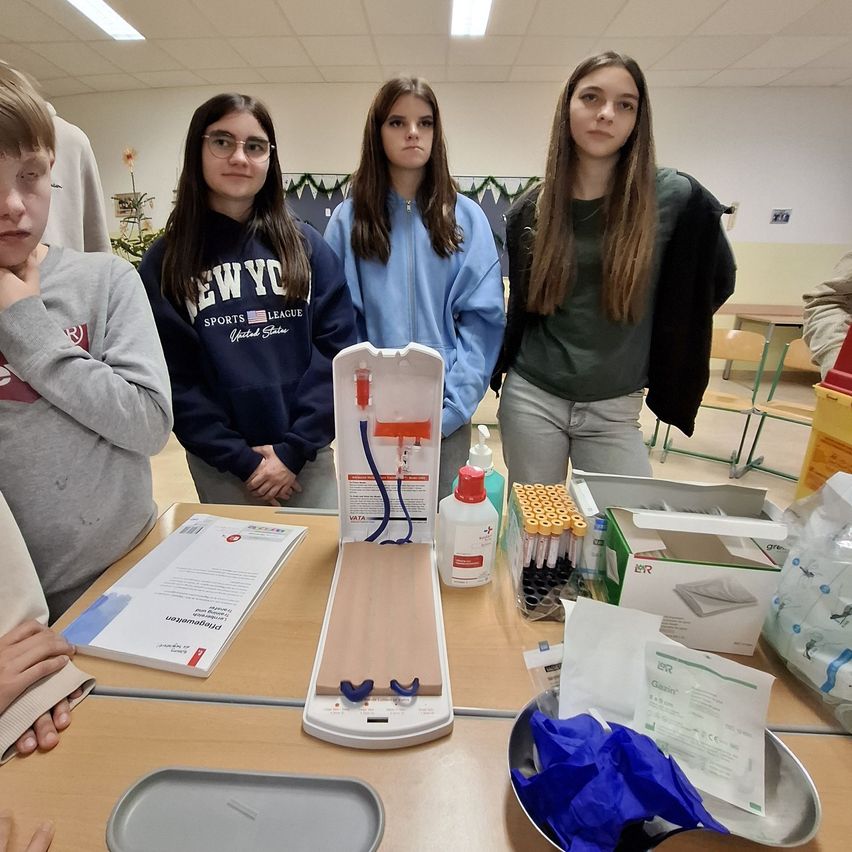 Vier junge Frauen stehen um einen Tisch mit verschiedenen medizinischen Geräten, einschließlich eines Blutentnahme-Kits. Eine Frau hält ein weißes Objekt, möglicherweise eine Blutprobe.