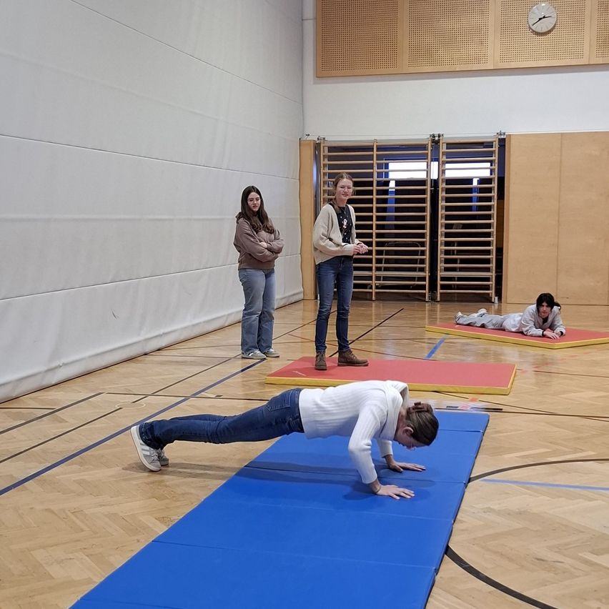 Eine Frau macht Liegestütze auf einer blauen Matte in einer Turnhalle. Zwei Frauen stehen hinter ihr, eine schaut sie an und die andere schaut etwas an. Eine weitere Frau liegt auf einer roten Matte. Eine Uhr ist an der Wand.