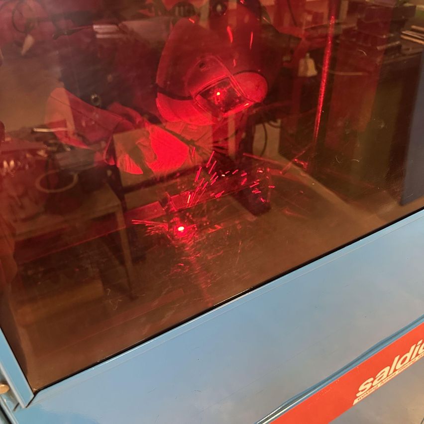 Ein Schweißer in Schutzkleidung arbeitet in einer Maschine, wobei ein rotes Leuchten vom Schweißen zu sehen ist. Die Maschine hat einen blauen und roten Rand und das Wort 'SALDIER' darauf.