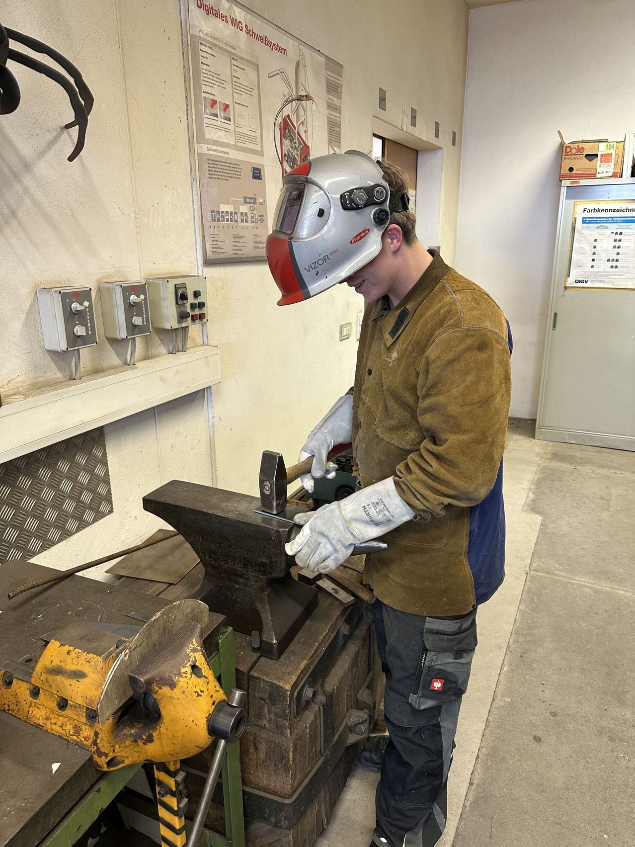 Ein Mann mit Schweißerhelm und Handschuhen arbeitet in einer Werkstatt an einem Amboss. Der Amboss steht auf einer Holzplatte.