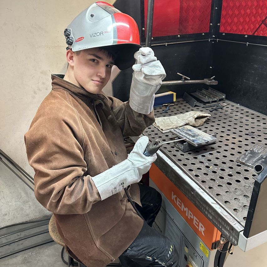 Ein junger Mann in Schweißerausrüstung arbeitet an einem Metallbearbeitungsprojekt an einer Metallbearbeitungsstation. Er hält ein Werkzeug in der Hand und lächelt.