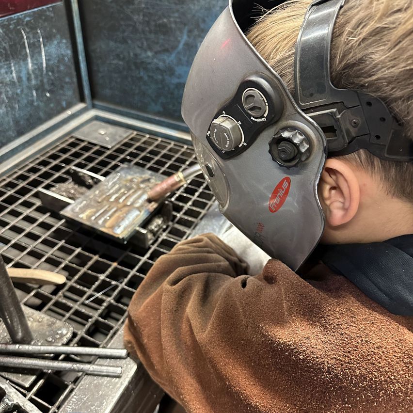 Ein junger Junge mit Schweißerhelm arbeitet an einer Metallbearbeitungsstation und konzentriert sich auf eine Metallplatte.