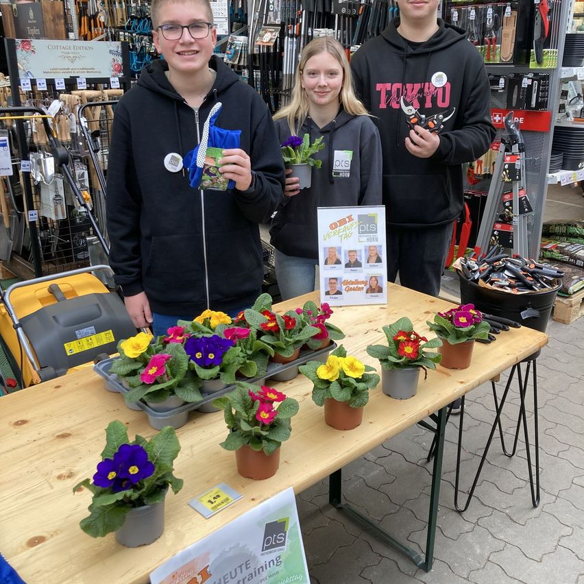 Drei Personen stehen hinter einem Tisch in einem Gartencenter und präsentieren verschiedene Topfpflanzen und Blumen, wobei Gartengeräte im Hintergrund sichtbar sind.