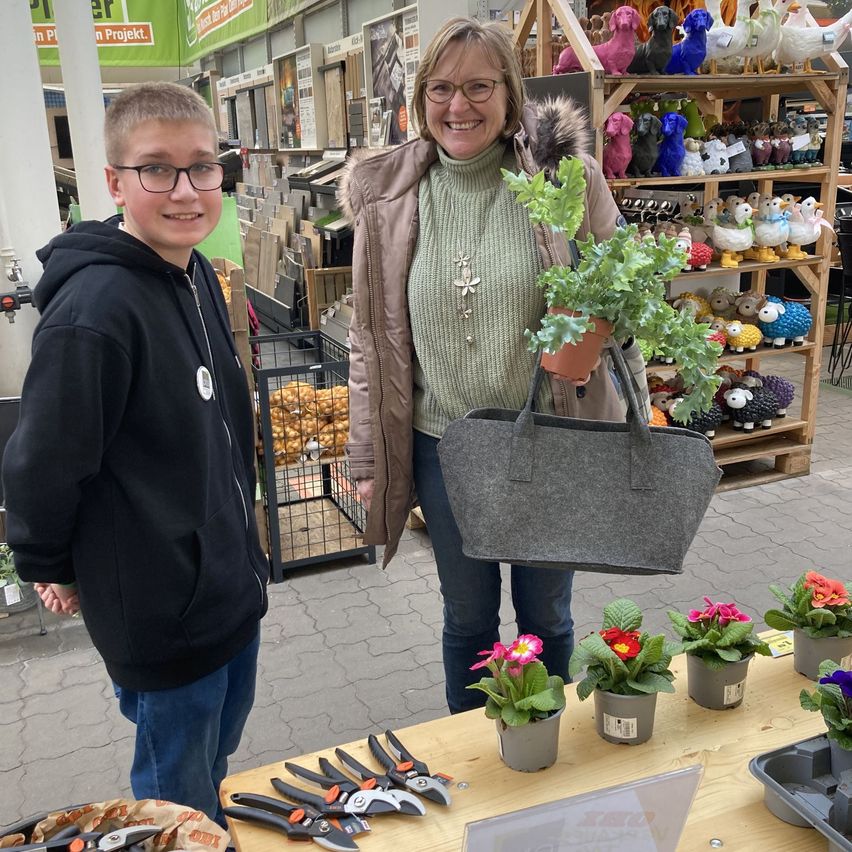 Eine Frau hält eine Pflanze und eine Handtasche in der Hand und steht hinter einem Tisch mit Topfblumen. Ein Junge mit Brille und Kapuzenpullover steht neben ihr. Im Hintergrund befinden sich Regale mit Plüschtieren.